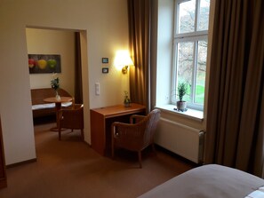 A hotel room with a bed, a desk, a chair, a window with curtains, a radiator, and a small potted plant. - Parkhotel del Mar (Sassnitz)