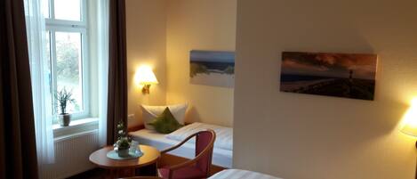 A hotel room with a bed, a chair, a small table with a plant, a lamp, and a painting on the wall.