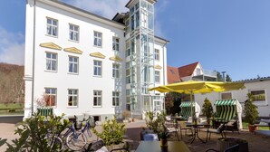 A white building with multiple windows and a glass staircase. There are outdoor tables and chairs with yellow umbrellas. - Parkhotel del Mar (Sassnitz)