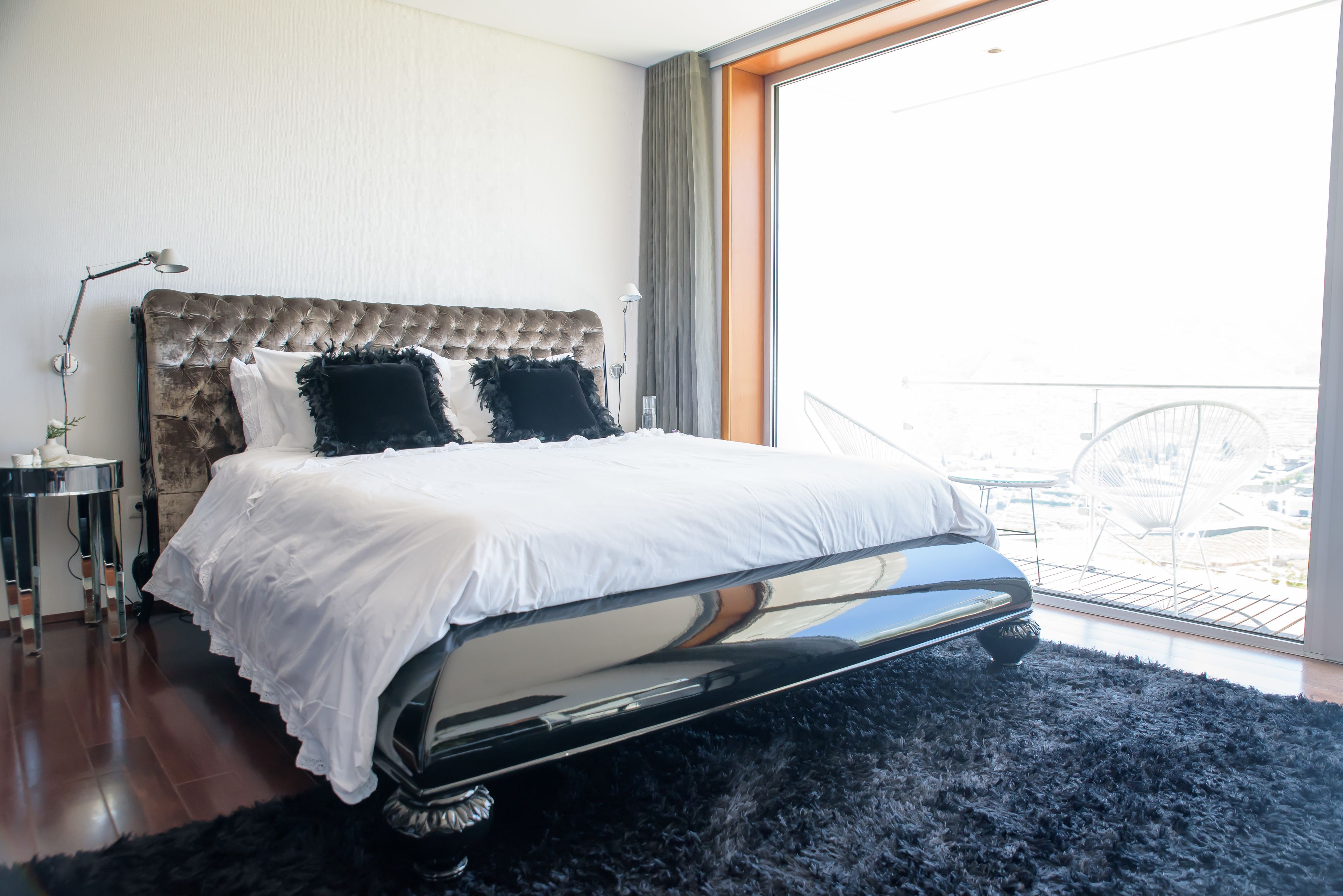 Une chambre avec une tête de lit capitonnée, une grande fenêtre donnant sur un beau paysage, un tapis noir et moelleux, et une table de chevet avec une lampe.