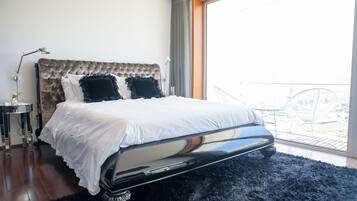 Une chambre avec une tête de lit capitonnée, une grande fenêtre donnant sur un beau paysage, un tapis noir et moelleux, et une table de chevet avec une lampe.