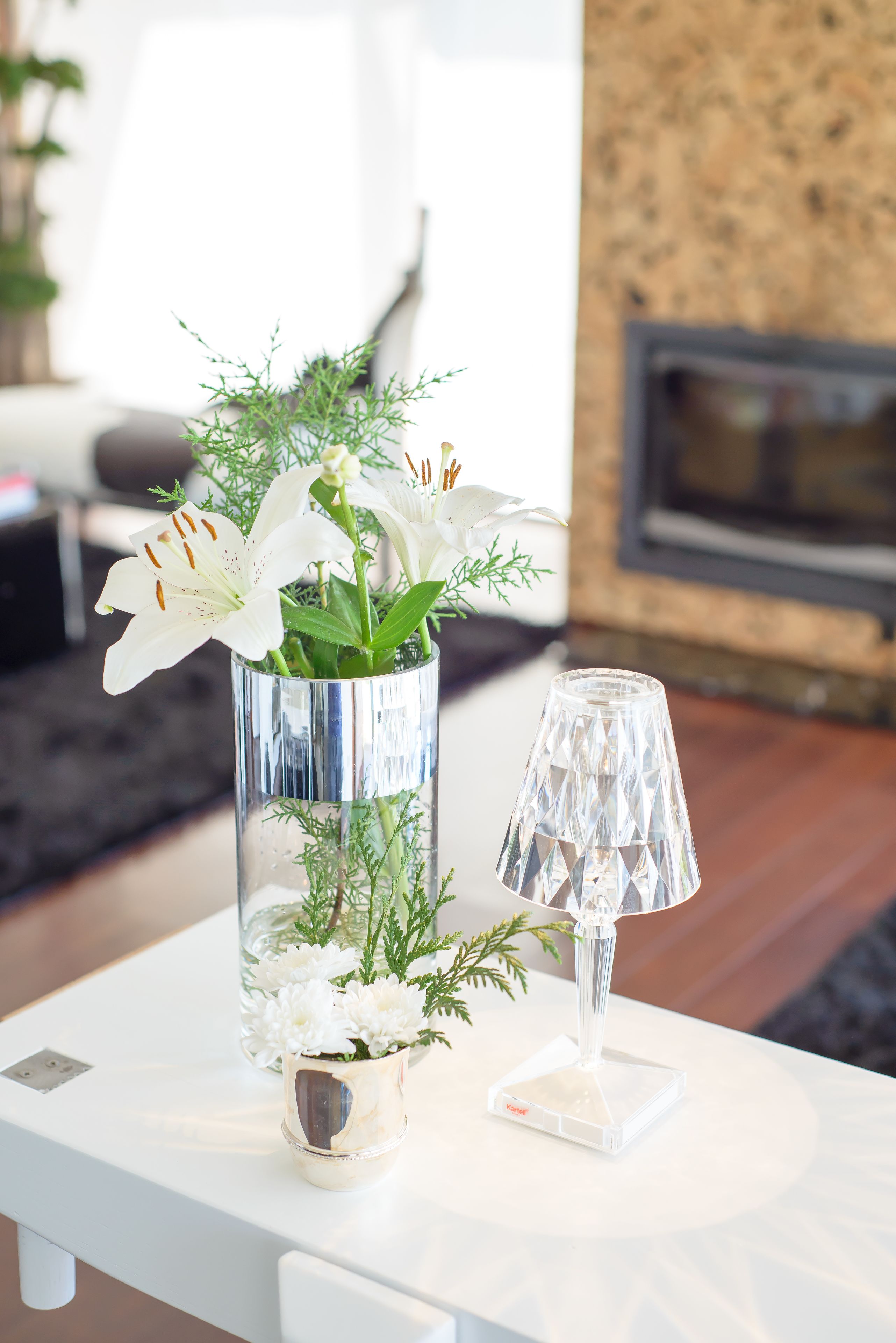 A white table with a glass vase containing white flowers and a clear glass lamp.