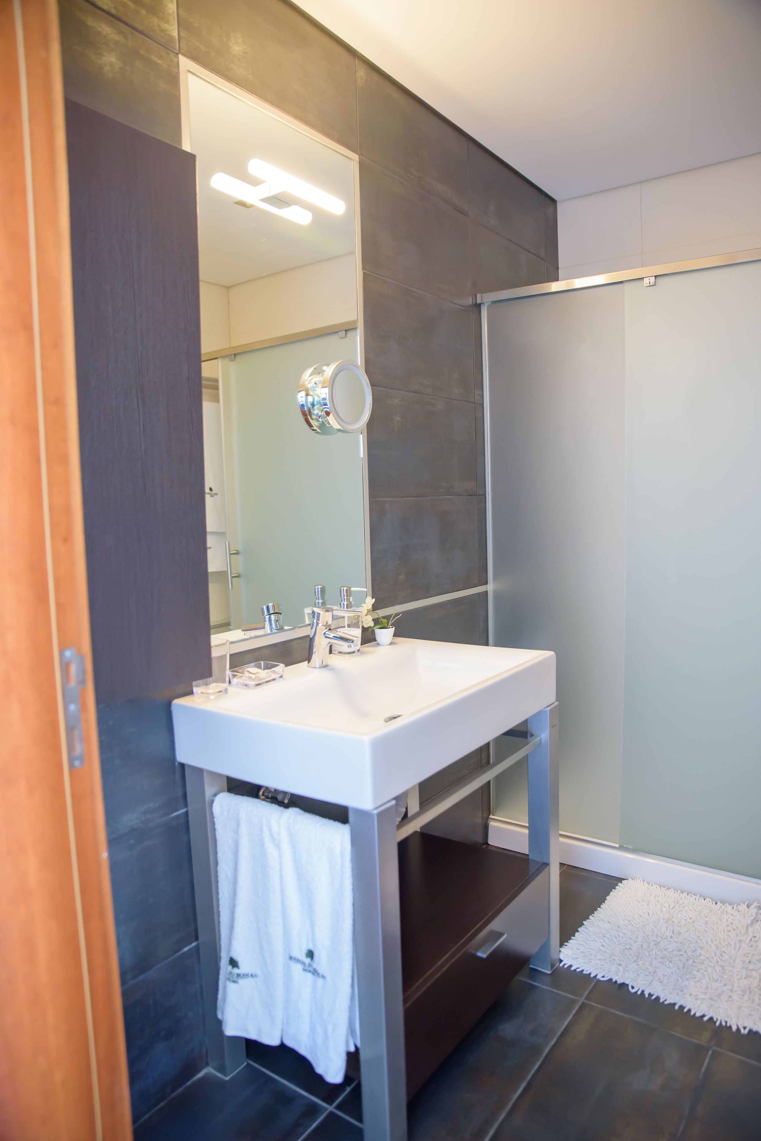 Un baño moderno con un espejo grande, lavamanos blanco y paredes cubiertas de azulejos oscuros.