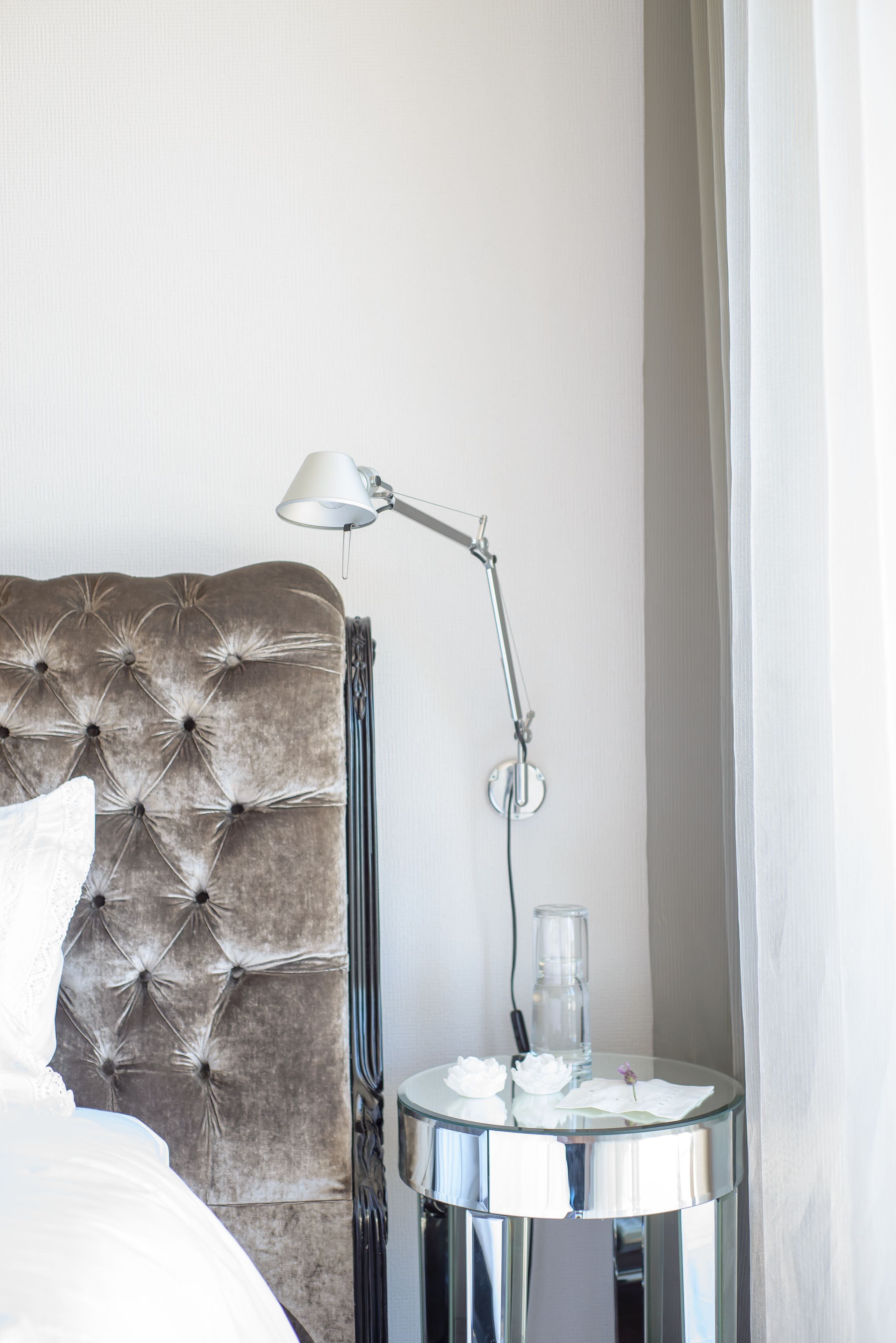 A neatly made bed with a tufted headboard, a bedside table with a lamp, and a glass vase with flowers.