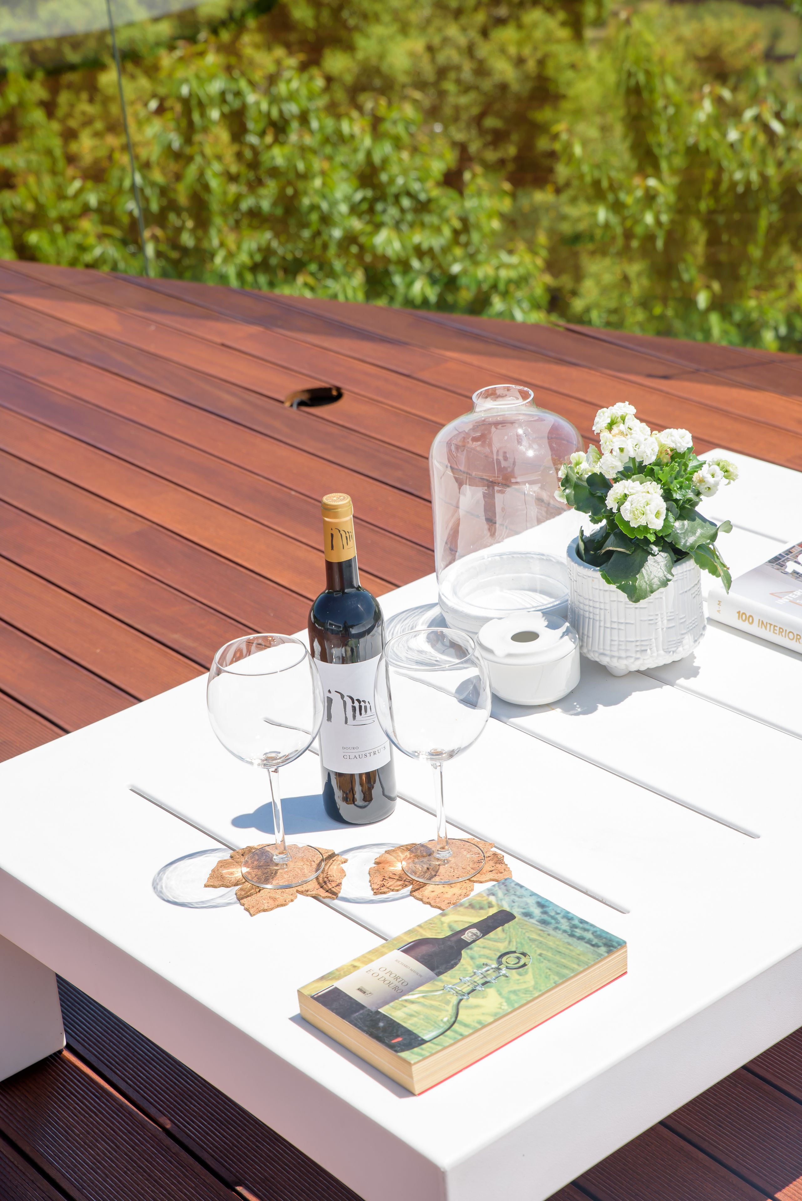 Mesa branca com garrafa de vinho, duas taças de vinho, um livro, um vaso com flores e um recipiente de vidro em um deck de madeira.