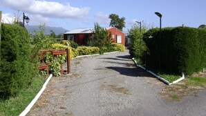 A gravel driveway leading to a red building with a white door, surrounded by greenery and a mountain in the background. - Cabañas Lago Azul (Puerto Varas)