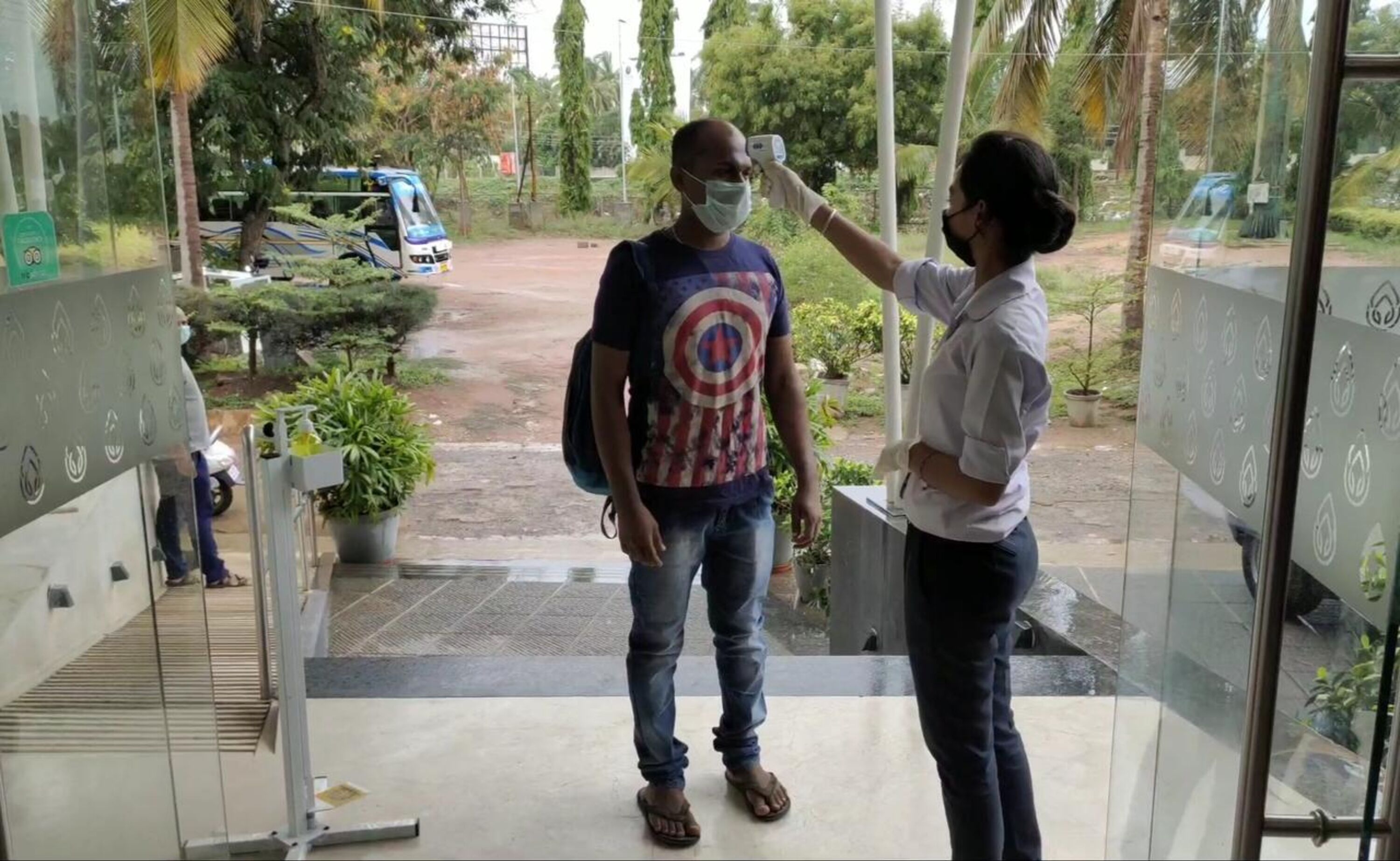 Une personne portant un masque et un t-shirt orné du logo de Captain America se tient dans un hall d’entrée, devant une porte vitrée, des plantes en pot et un véhicule garé à l’extérieur.