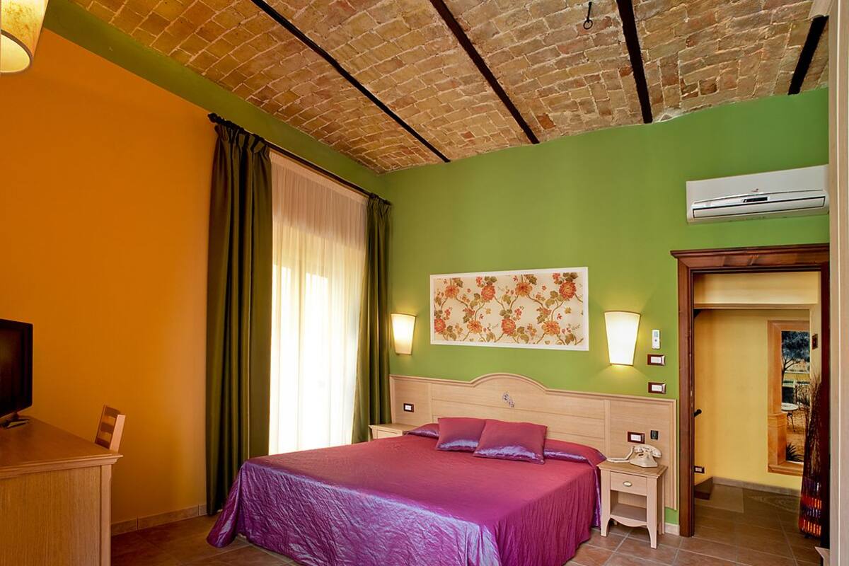 A bedroom with a brick ceiling, green walls, a bed with a purple bedspread, a nightstand, a lamp, and a TV on a wooden desk.