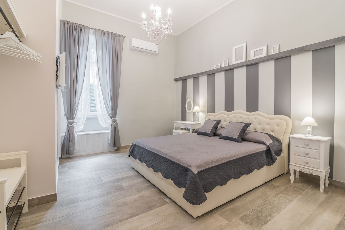 Un dormitorio con una cama grande, mesitas de noche, un candelabro y una ventana con cortinas.