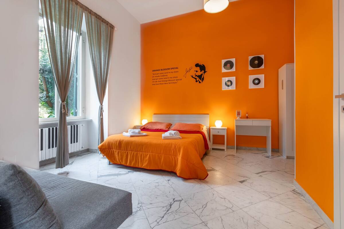 A hotel room with an orange accent wall, a bed with orange bedding, a bedside table with a lamp, and a window with curtains.
