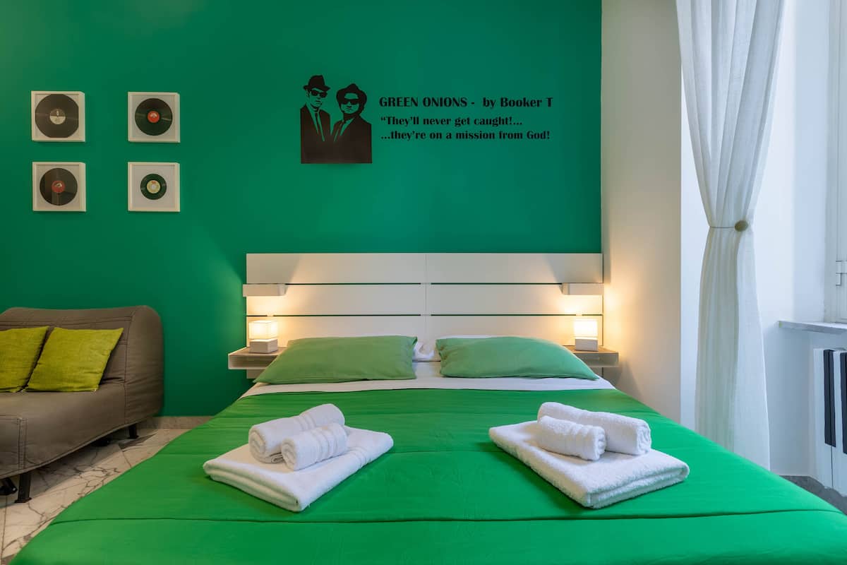 A hotel room with a green bedspread, two pillows, a headboard, and a wall with a poster and four framed pictures.