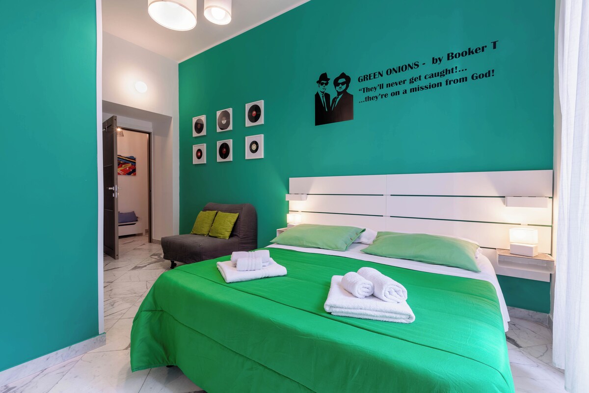 Quarto de hotel moderno com decoração em tons de verde, destacando-se uma cama grande com roupa de cama verde, um sofá cinzento e uma parede com quadros e um pôster de greenions.