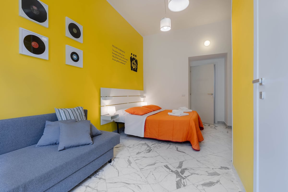 A room with a yellow accent wall, a bed with an orange bedspread, a blue sofa, and a white headboard.
