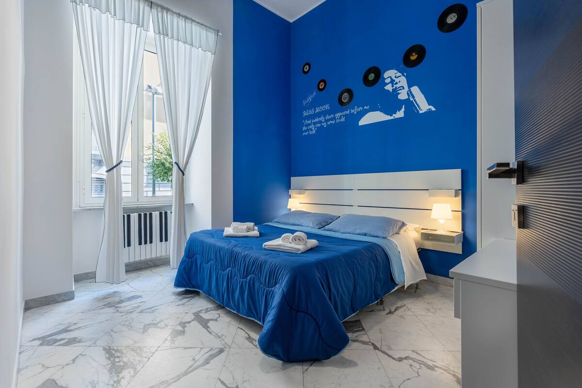 A bedroom with a blue bedspread, white curtains, a wall mural, and a marble floor.
