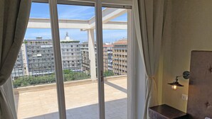 A bedroom with a large sliding glass door leading to a balcony. The room has a wooden headboard, a bedside table with a lamp, and a bed with a white pillow.