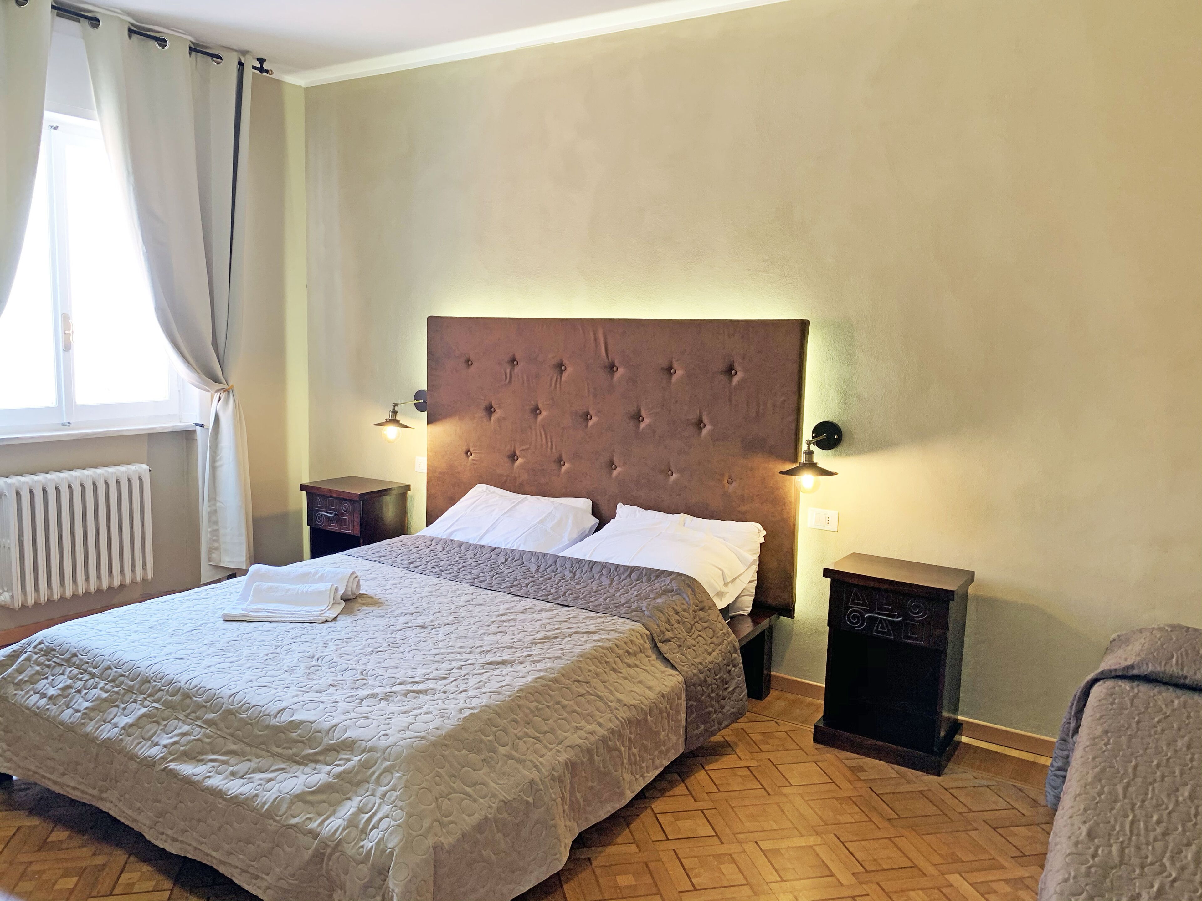 A hotel room with a large bed, two bedside tables, a radiator, and a window with curtains.