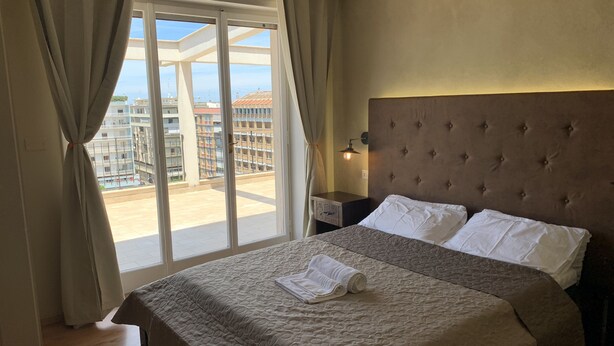 A hotel room with a large bed, a nightstand, a lamp, and a view of buildings through a large window.