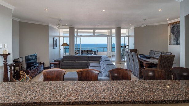 A spacious living room with a large sectional sofa, a dining area with chairs, a kitchen with granite countertops, and a view of the ocean through large windows. - 702 Oyster Rock (Umhlanga)