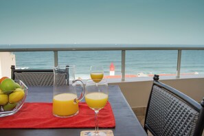 Une table avec une carafe et deux verres de jus d’orange, un saladier de fruits, et une vue sur l’océan avec un phare au loin.