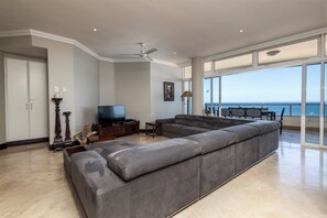 Un salon doté d’un grand canapé d’angle gris, d’un téléviseur à écran plat, d’un ventilateur de plafond et offrant une vue sur l’océan grâce à de grandes fenêtres.