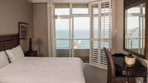 A hotel room with a bed, a chair, a desk, a lamp, a vase with flowers, and a large window with a view of the ocean and a lighthouse. - 702 Oyster Rock (Umhlanga)