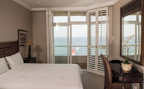 Une chambre d’hôtel comprenant un lit, une chaise, un bureau, une lampe, un vase avec des fleurs et une grande fenêtre donnant sur l’océan et un phare.