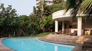A swimming pool with lounge chairs and a covered seating area, surrounded by lush greenery and a tall building in the background. - 702 Oyster Rock (Umhlanga)