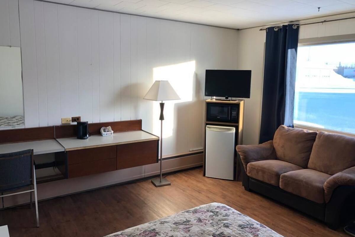 Quarto de hotel com piso de madeira, sofá, escrivaninha com luminária, televisão e uma pequena geladeira.