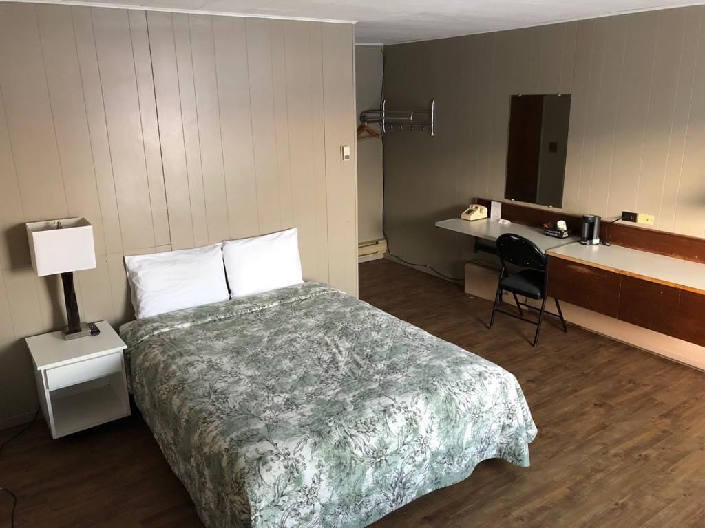 A hotel room with a bed, a desk, a chair, a nightstand, a lamp, a mirror, and a telephone.
