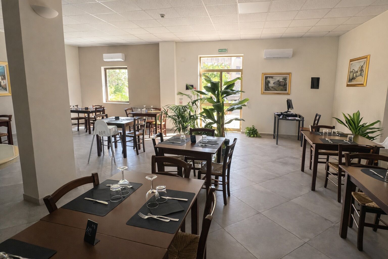 Un comedor amplio con varias mesas y sillas, ventanas grandes que permiten la luz natural y plantas de interior.