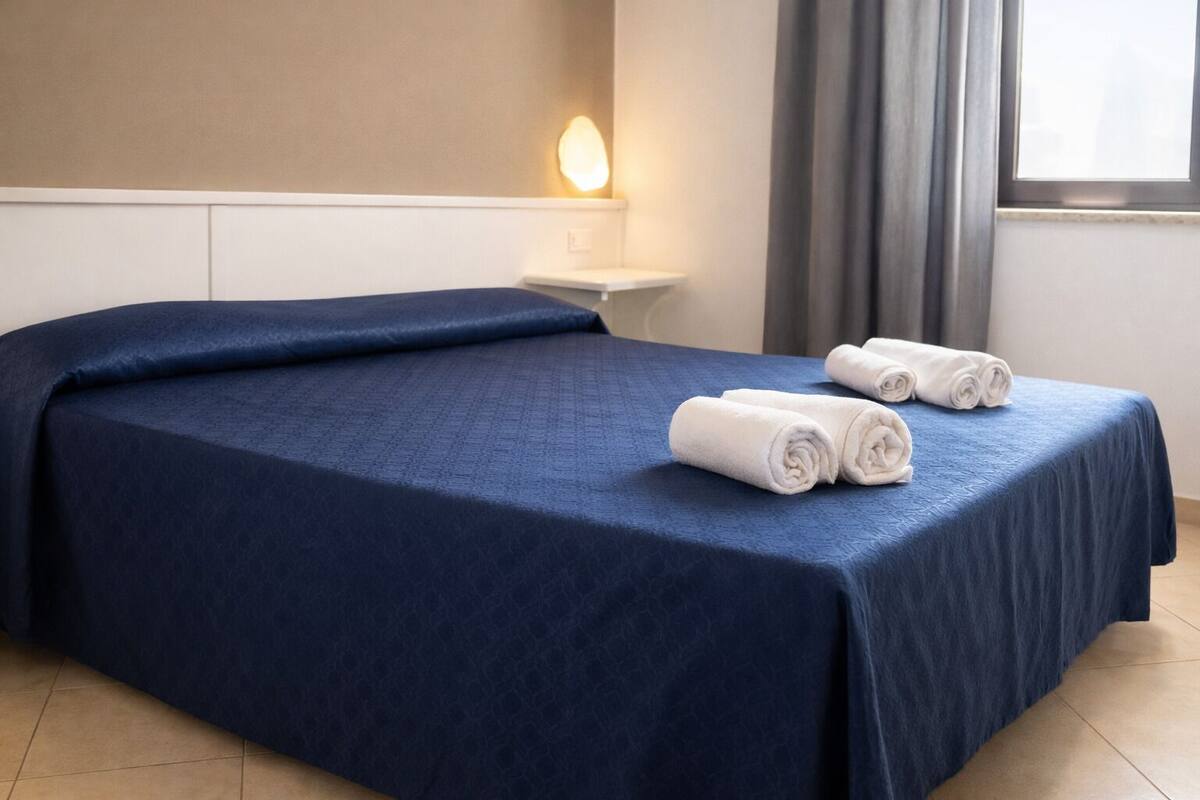 A hotel room with a large bed, a bedside table, a wall-mounted light, and a window with curtains.