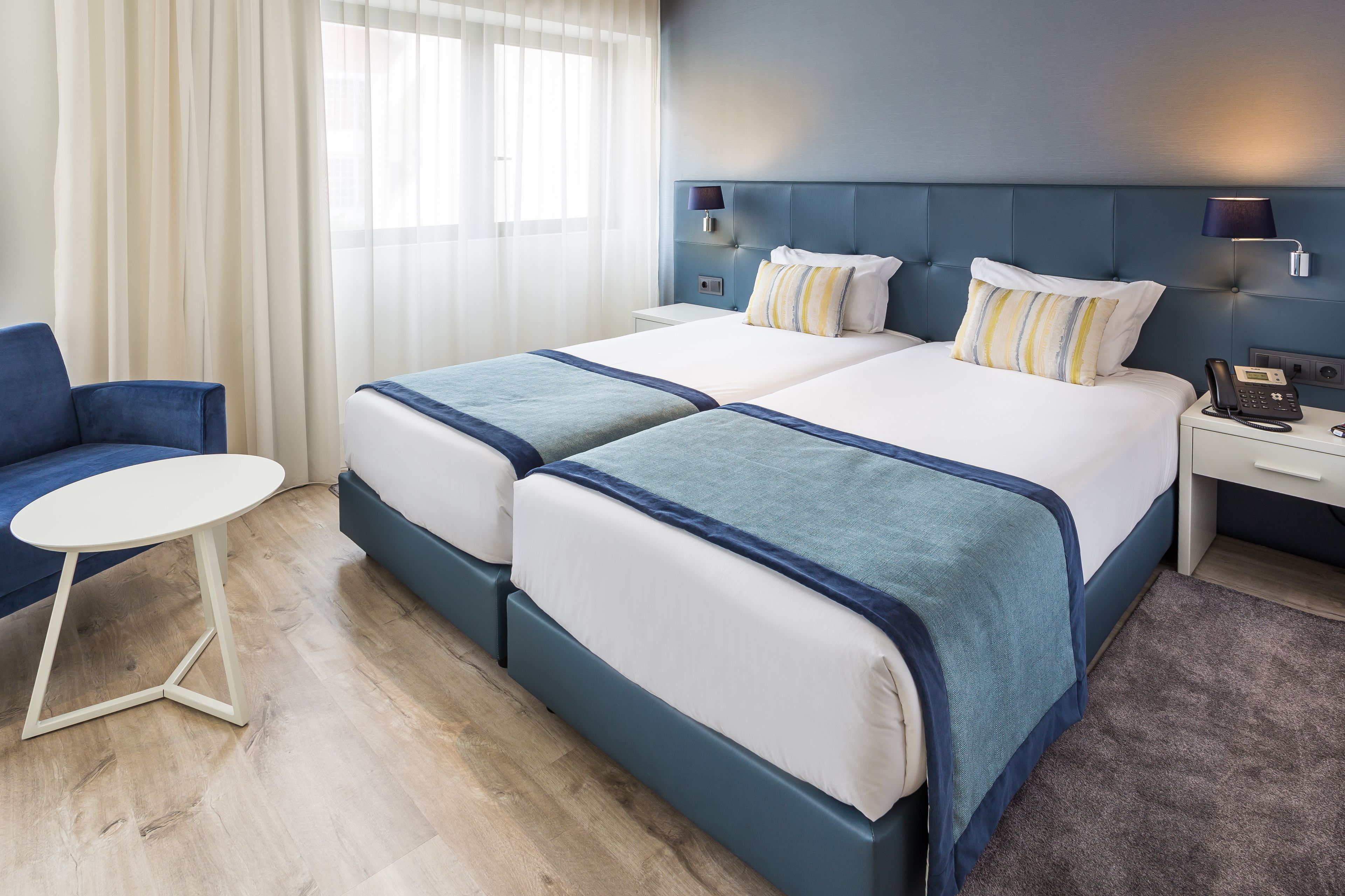 A hotel room with two beds, a small table, a chair, and a nightstand with a phone.