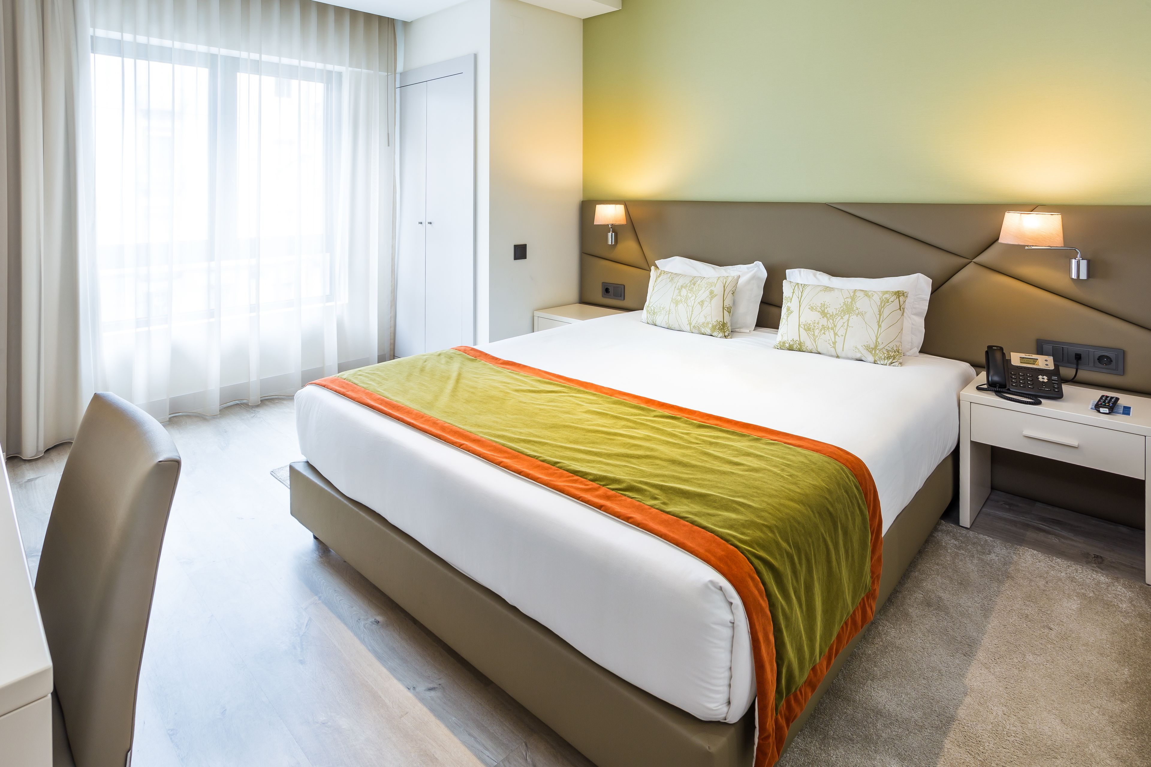 Une chambre d’hôtel avec un grand lit, un bureau et une chaise. Il y a deux tables de chevet avec des lampes et un téléphone.