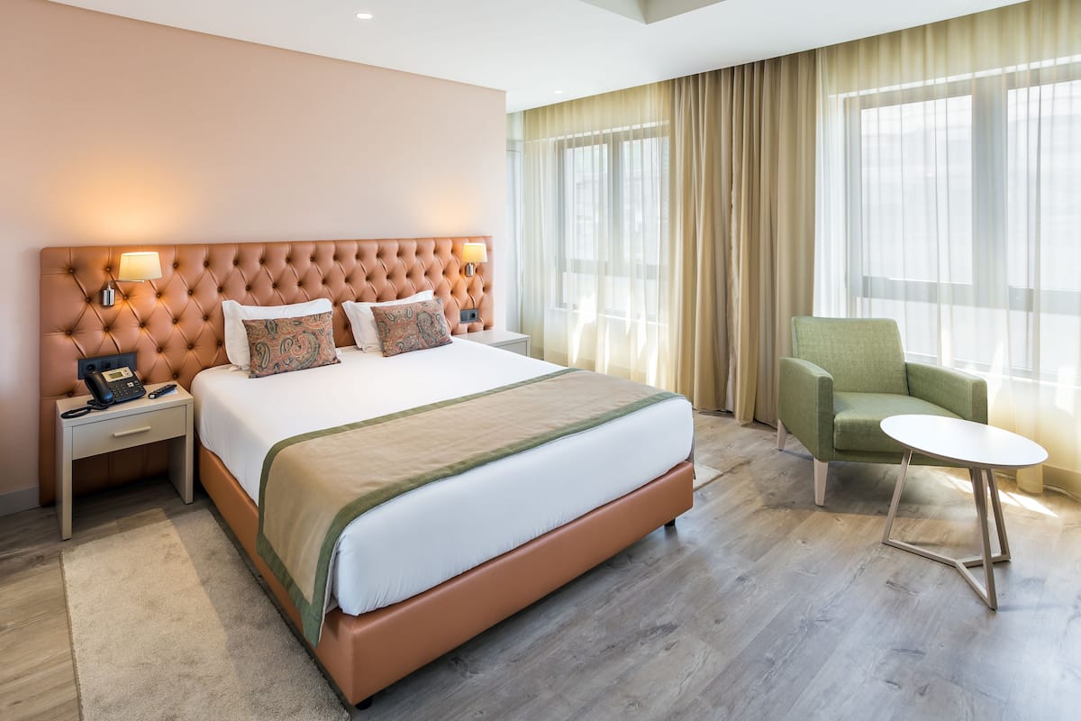 Een hotelkamer met een groot bed, een groene fauteuil, een klein rond tafeltje en een nachtkastje met een lamp en telefoon.