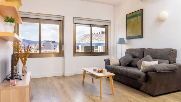 A modern living room with a grey sofa, wooden coffee table, and large windows offering a city view. - Apartamentos Mayerling Bisbe Urquinaona (Barcelona)