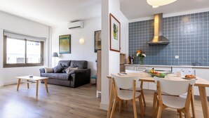 A modern living room with a grey sofa, a wooden dining table with chairs, and a kitchen area with blue tiles and a stainless steel range hood. - Apartamentos Mayerling Bisbe Urquinaona (Barcelona)