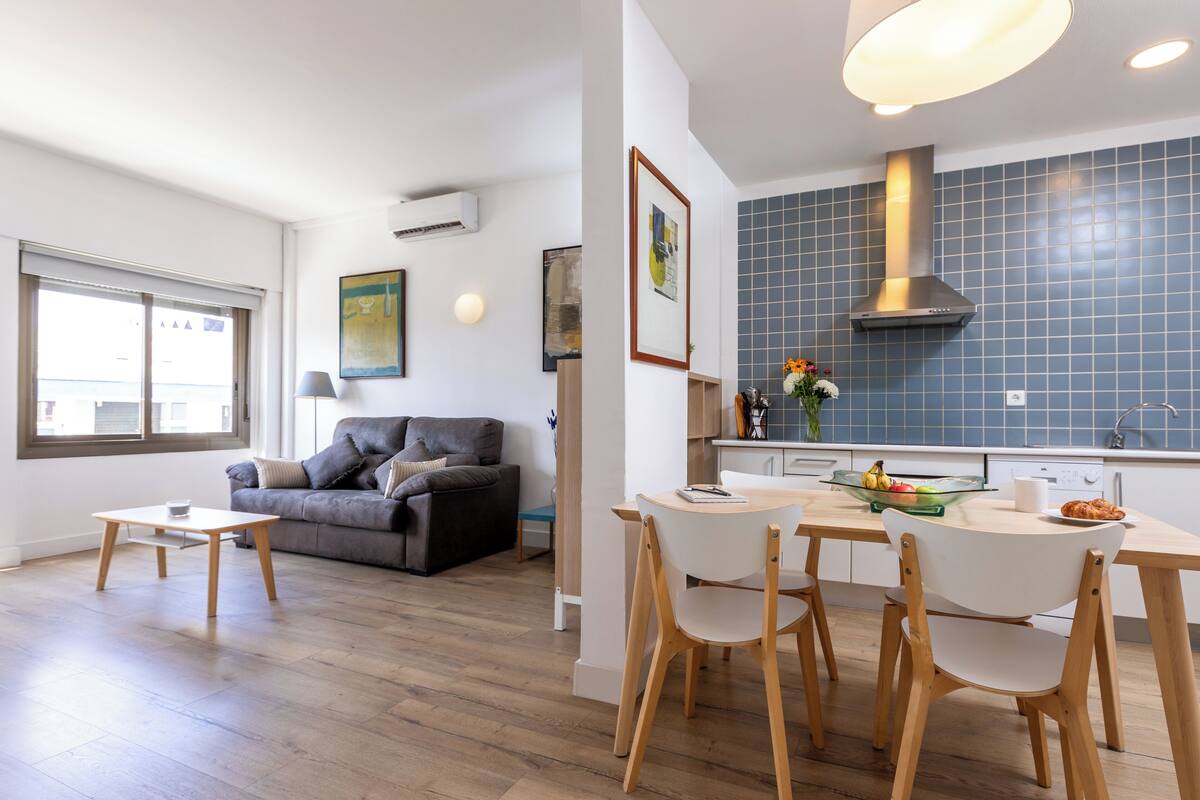 A modern living room with a grey sofa, a wooden dining table with chairs, and a kitchen area with blue tiles and a stainless steel range hood.