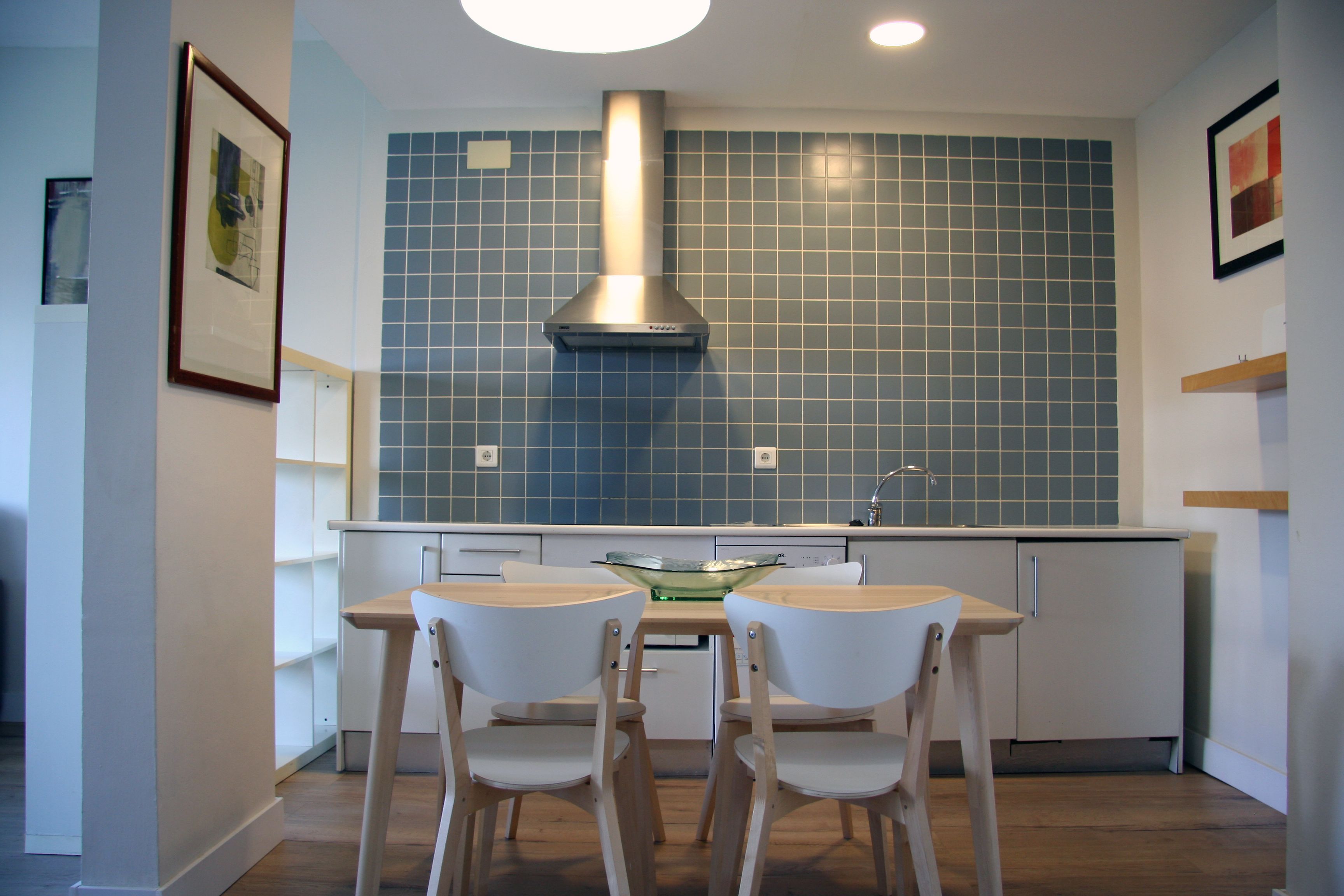 Een moderne keuken met blauwe tegels, een roestvrijstalen afzuigkap, een glazen kom op het aanrecht en twee houten stoelen.