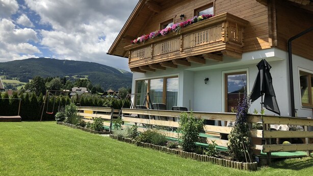 Une maison en bois à deux étages, dotée d’un balcon orné de fleurs, d’une clôture en bois et d’une cour gazonnée.