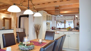 Un coin repas avec une table en bois, des chaises et un lustre. On aperçoit au fond une cuisine avec des meubles blancs et une fenêtre ornée de fleurs.