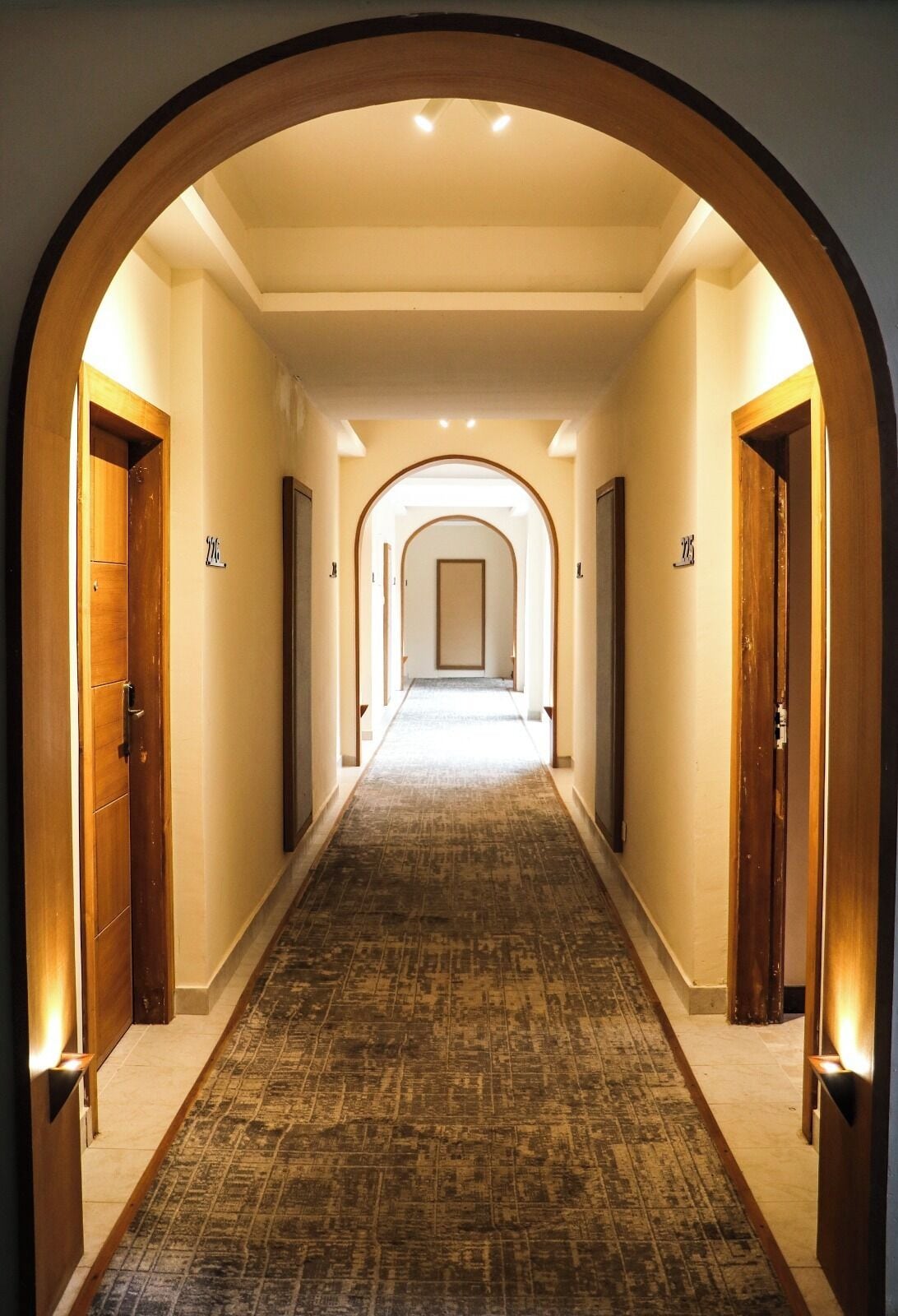 A hotel hallway with a carpeted floor, wooden doors, and a central archway.