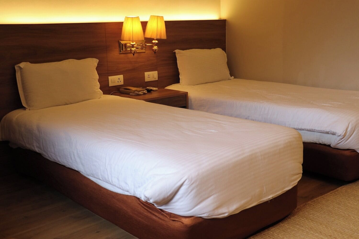 Una habitación de hotel con dos camas, cada una con ropa blanca y cabeceros de madera. Hay dos lámparas fijadas a los cabeceros.