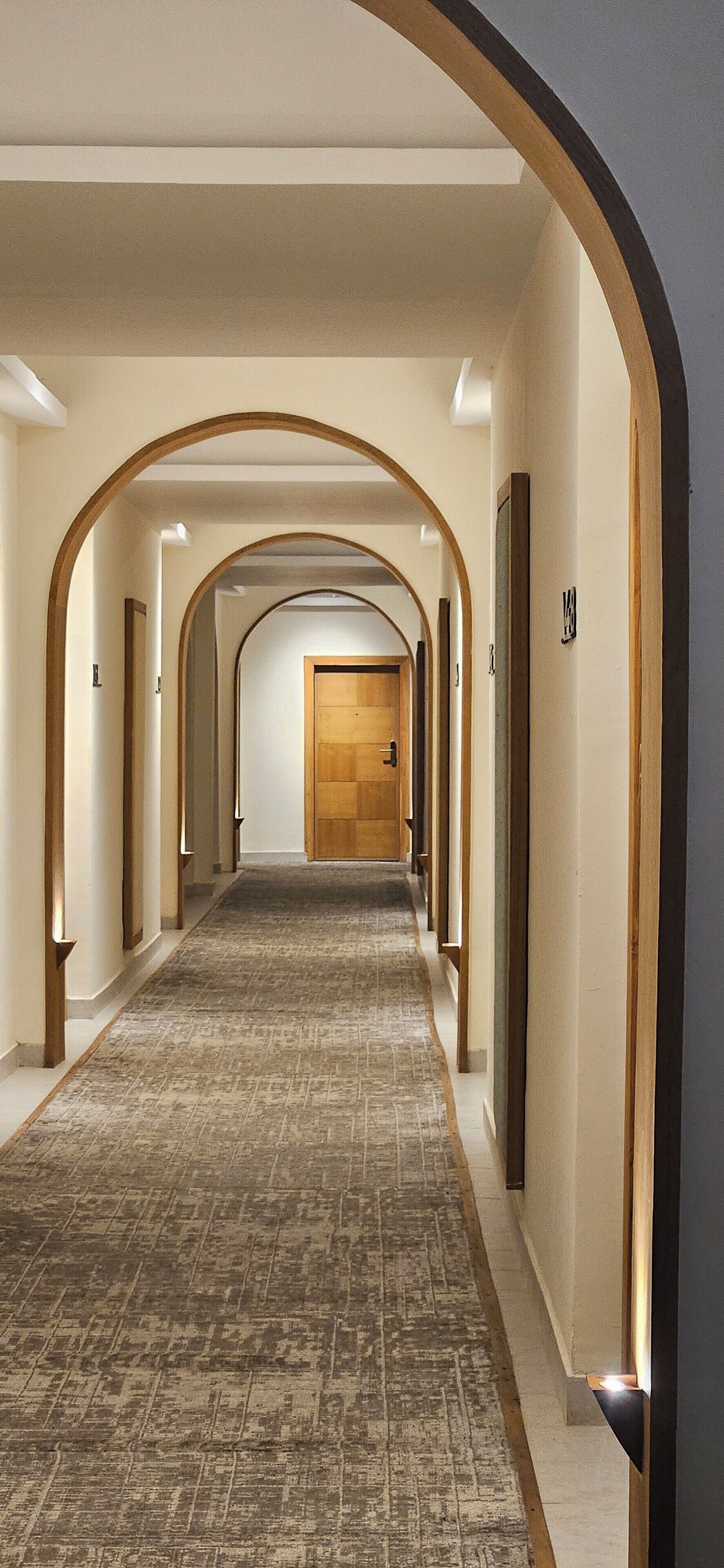 Un couloir d’hôtel avec un sol recouvert de moquette, des encadrements de porte en bois et une série de portes le long du couloir.