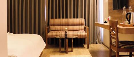 A hotel room with a bed, a sofa, a small table with a chair, a TV mounted on the wall, and a curtain backdrop.
