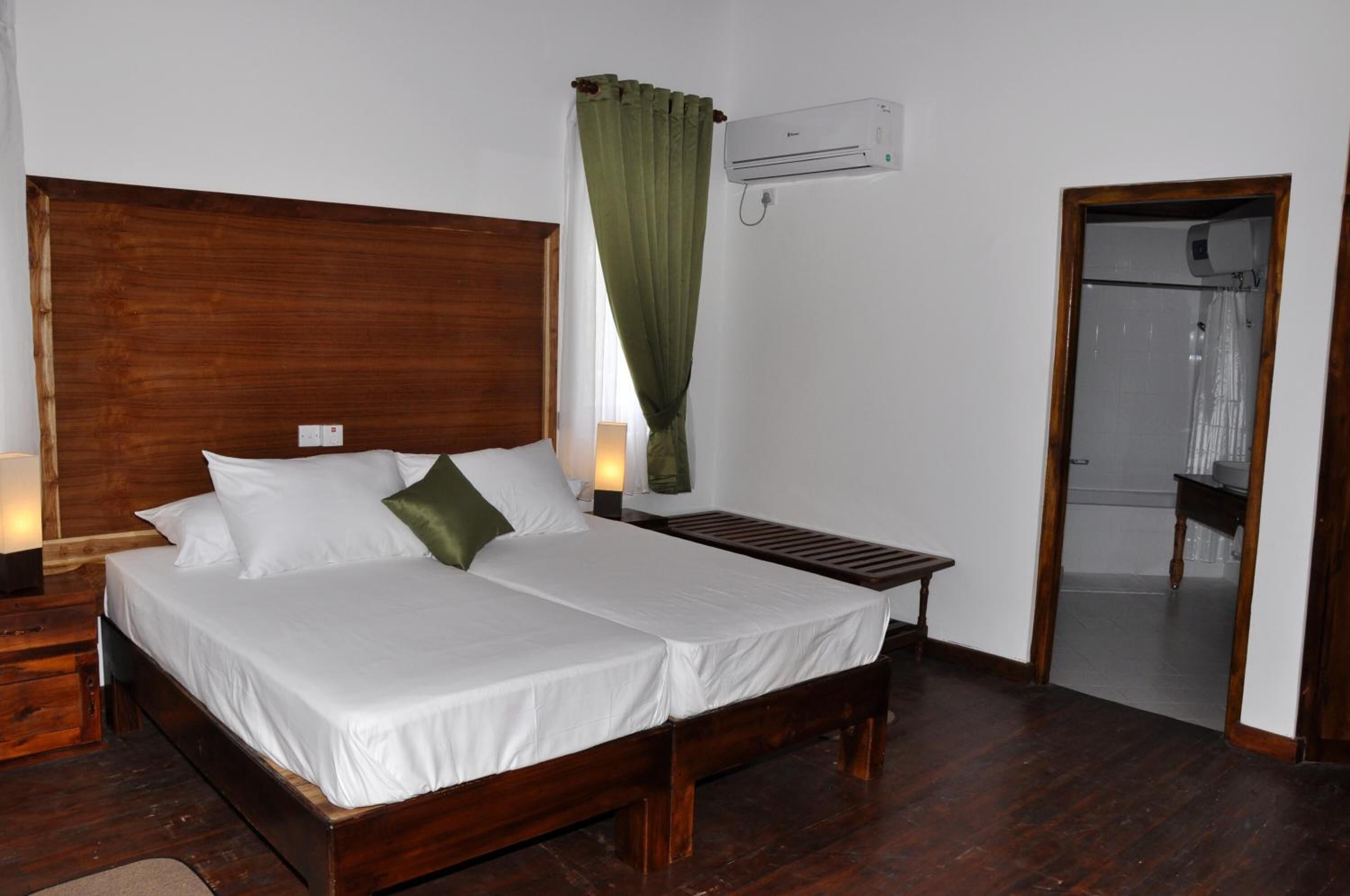 A double bed with white linens, a wooden headboard, a bedside table with a lamp, and a green curtain.