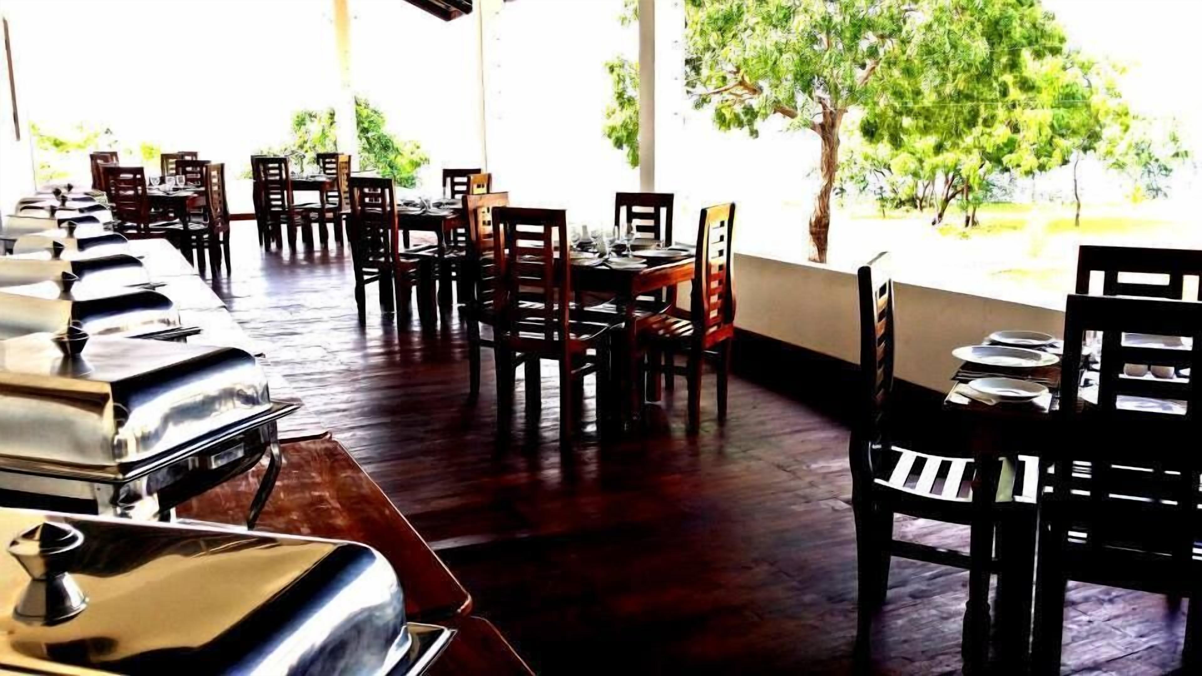 A restaurant with wooden tables and chairs, a long counter with a grill, and large windows with a view of trees.