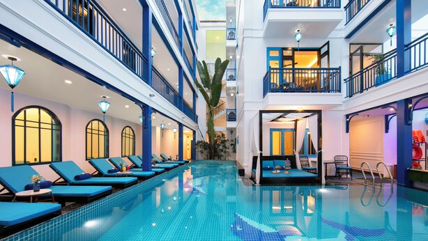A hotel interior with a swimming pool, lounge chairs, and a staircase leading to upper floors. - Silk River Hoi An Retreat (Da Nang)