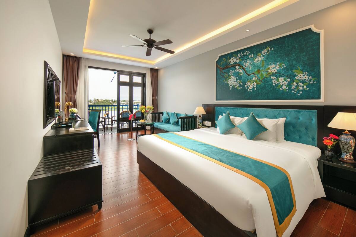 Habitación de hotel con una cama grande, ventilador de techo, un cuadro y balcón con mobiliario exterior.