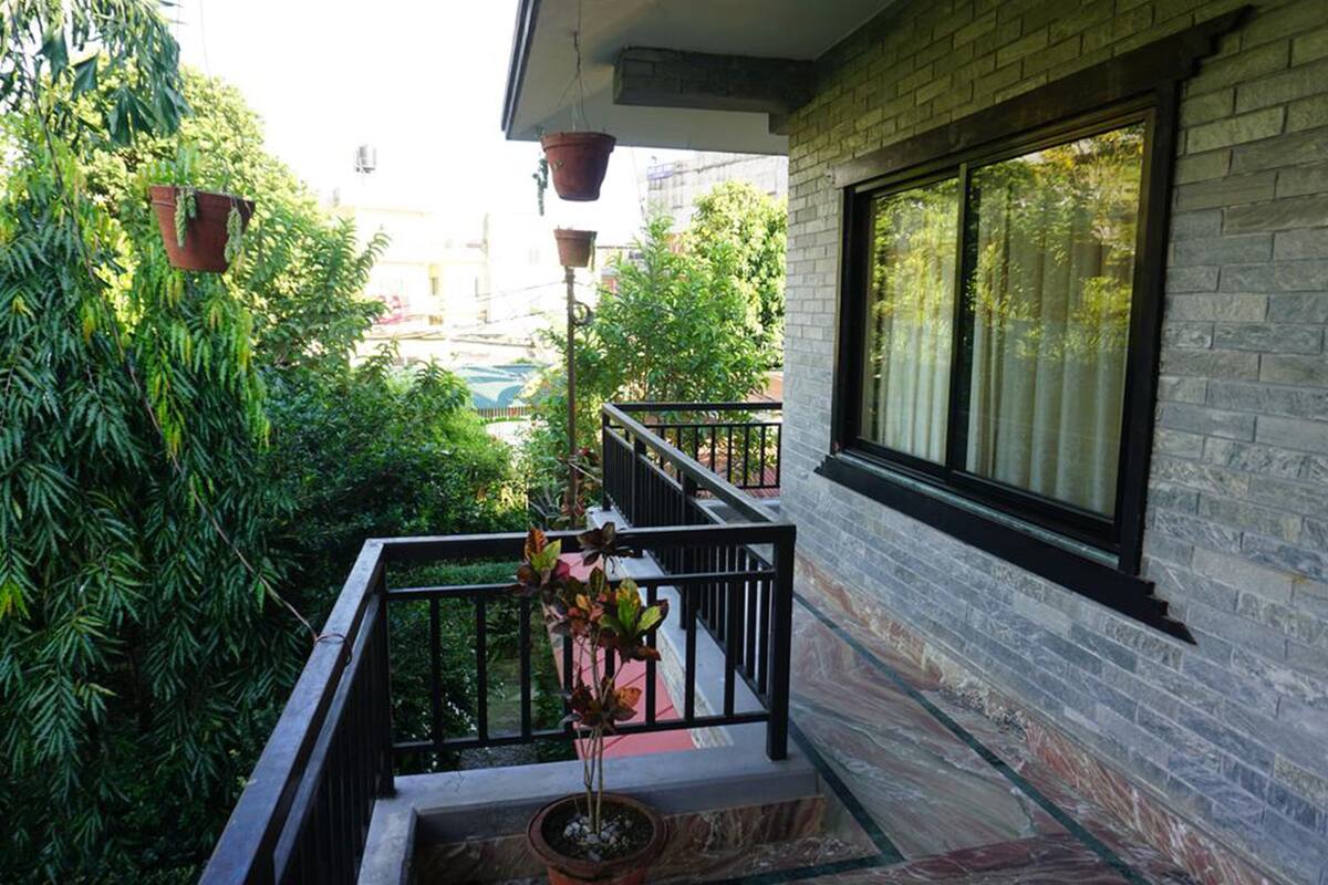 A balcony with a potted plant, a window with a view of greenery, and a brick wall.