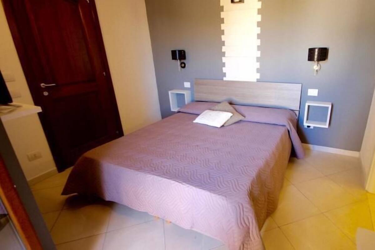 A bedroom with a bed, a nightstand, a wall-mounted lamp, and a door.